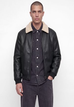 Un mannequin masculin se tient devant un fond blanc, vêtu d’une veste en cuir noire à col en peau lainée couleur crème et d’un jean foncé.