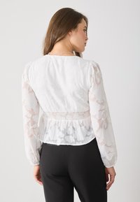 White blouse with sheer long sleeves, floral texture, and smocked waist detail. Paired with black trousers, displayed from the back.