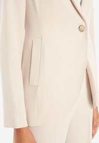 Beige blazer i mjukt tyg, med enspärrad knäppning, sidoficka och rundad nederkant, samt något skräddarsydda ärmar.