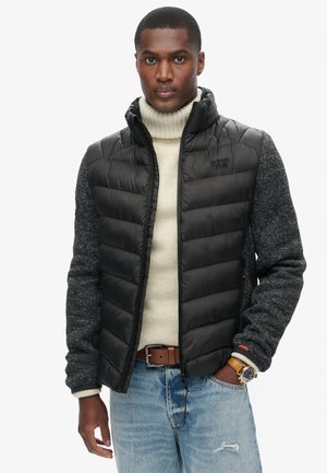Giacca nera imbottita con maniche a maglione grigie, collo alto e chiusura con zip. Indossata su un dolcevita beige. Visibili jeans blu e un orologio.
