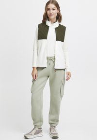 Quiltad olivgrön väst över en vit långärmad skjorta, kombinerat med ljusgröna cargobyxor och beige sneakers. Har dragsko i midjan och fickor.