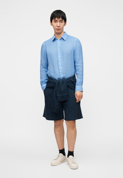 Ανοιχτό μπλε λινό πουκάμισο με γιακά με κουμπιά, σε συνδυασμό με σκούρα ναυτικά σορτς και λευκά αθλητικά παπούτσια. Το πουκάμισο έχει μανίκια που είναι γυρισμένα και χαλαρή εφαρμογή.