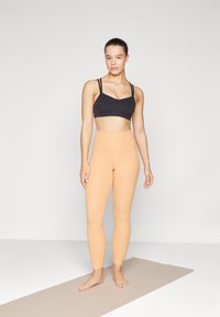 Reggiseno sportivo nero con due spalline sottili, abbinato a leggings a vita alta di un arancione chiaro. Texture liscia, design aderente, branding minimalista.