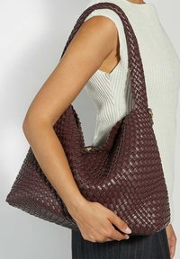 Woven burgundy shoulder bag with a soft texture, round shape, and a wide strap, featuring a gold hardware accent.