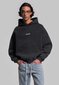 Felpa nera oversize con tasca frontale e testo bianco "WRSTBHVVR". Indossata sopra jeans azzurri chiari, che evidenziano un taglio rilassato e un design casual.