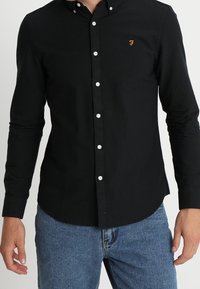 Hombre vestido con una camisa negra ajustada de botones con una pequeña "F" bordada en naranja en el pecho y jeans azules, de pie con los brazos relajados.