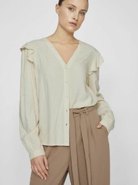 Lys beige bluse med V-hals og knaplukning foran, med flæset skuldre og lange ærmer. Glat tekstur.