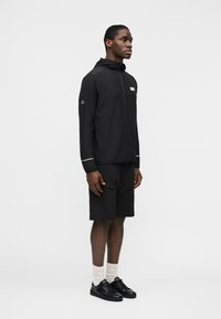 Άνδρας στέκεται με κατεύθυνση προς τα δεξιά, φορώντας μαύρο μπουφάν με κουκούλα και φερμουάρ, μαύρα σορτς μέχρι το γόνατο, άσπρες κάλτσες και μαύρα αθλητικά παπούτσια, σε λευκό απλό φόντο.