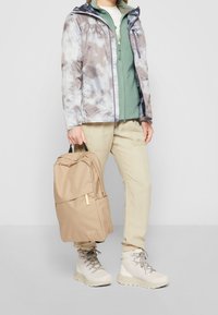 Lichte, tie-dye jas in grijs en wit, gelaagd over een groene fleece, gecombineerd met beige broek en een tan rugzak, en crèmekleurige wandelschoenen.