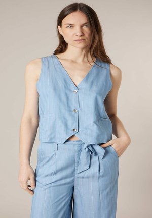 Vrouw draagt mouwloos lichtblauw vest met knopen en bijpassende hoge taillebroek met ceintuur, hand in zak, neutrale achtergrond.