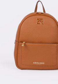 Zaino in pelle marrone dalla texture texturizzata e dalla forma arrotondata, con un logo dorato, dettagli con cerniere dorate e una tasca frontale.