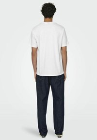 T-shirt bianco in cotone con maniche corte, visto da dietro. Indossato con jeans in denim blu scuro a vestibilità ampia con dettagli di cucitura visibili.