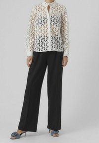Chemise en dentelle boutonnée avec motif floral en blanc et beige, associée à un pantalon large noir et des sandales à talons bleues ornées d'un accent floral.