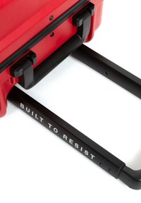 Red suitcase with a textured exterior, black plastic handles, and a sturdy retractable handle featuring the text "BUILT TO RESIST."