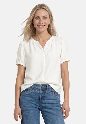 Lächelnde Frau mit blondem Haar, die eine weiße kurzärmelige Bluse und blaue Jeans trägt, vor einem schlichten weißen Hintergrund stehend.