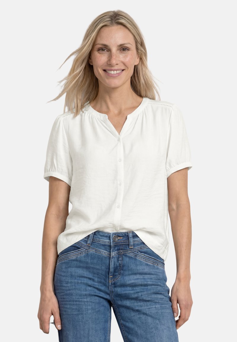 Femme souriante aux cheveux blonds portant un chemisier blanc à manches courtes et un jean bleu, debout devant un fond blanc uni.