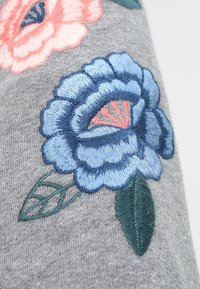 Grått tyg med blå och rosa broderade blommor med detaljerade kronblad och gröna blad, som visar en strukturerad design.