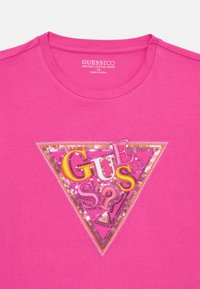 Tricou roz din bumbac organic, cu o aplicație triunghiulară din paiete având textul "GUESS" în aur și accente multicolore. Decolteu rotund.