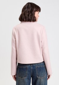 Giacca corta rosa chiaro realizzata in tessuto morbido con colletto, caratterizzata da maniche lunghe e una texture liscia. Abbinata a jeans scuri.