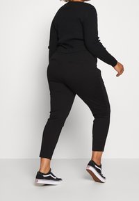 Pantaloni neri aderenti in un tessuto testurizzato, con tasche anteriori e posteriori e un design a gamba affusolata, abbinati a sneaker nere.