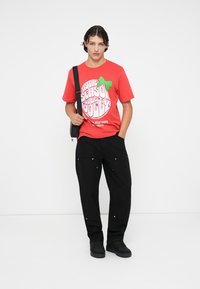 Tricou roșu cu un grafic circular pe care scrie "Este sezonul să fim veseli", asociat cu o pereche de pantaloni negri largi, cu buzunare frontale și accente metalice.