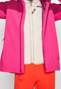 Roze en kastanjebruine lichtgewicht jas over een crème fleece binnenlaag, met een ritssluiting en oranje broeken. Geconfigureerde materialen overal.