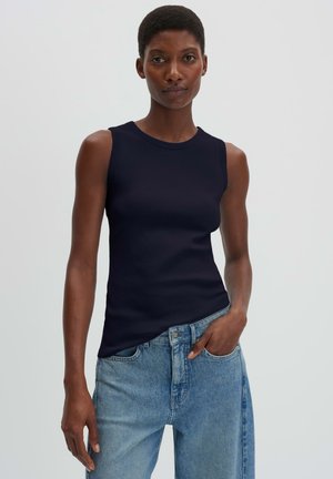Femme portant un haut noir sans manches et un jean bleu ample avec une main dans la poche, devant un fond clair uni.