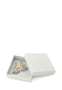 Goudkleurige oorbellen met bloemmotief en blauw druppelaccent, tentoongesteld in een lichtgrijze vierkante doos met een subtiel reliëflogo.