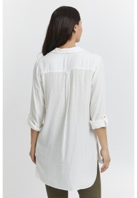 Weiße Bluse mit einem strukturierten Muster, langen, hochgerollten Ärmeln und einem Rückenpasse. Sie hat einen längeren Rückensaum und Knopfdetails an den Manschetten.