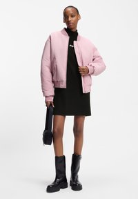 Roze bomberjack met geribbelde manchetten, zwarte coltrui-jurk en zwarte kniehoge laarzen. Model draagt een zwarte crossbodytas.