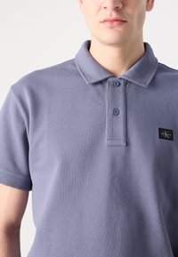 Polo en tissu bleu clair avec une finition texturée, présentant un col, deux boutons et un petit écusson logo noir sur la poitrine.