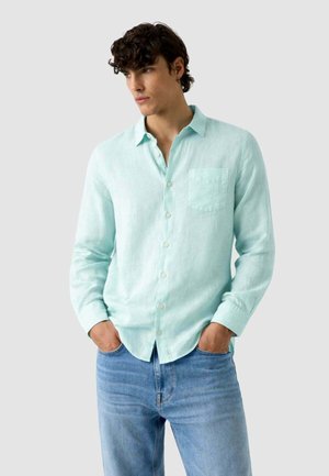 Giovane uomo con capelli scuri e ricci che indossa una camicia di lino azzurro chiaro abbottonata e jeans blu, in piedi con le mani in tasca.