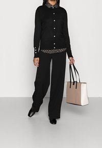 Black button-up cardigan with decorative buttons, layered over a patterned shirt. Wide black trousers and a beige tote bag with black accents.