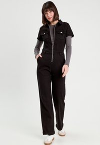 Tuta nera con maniche corte, caratterizzata da una cerniera frontale e due tasche sul petto. Abbinata a una maglietta grigia a maniche lunghe sottostante.