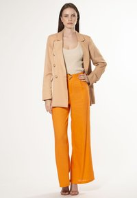 Beige dubbelknoop blazer, crèmekleurige tanktop, en levendig oranje wijde broek met een cirkelvormige riemgesp en een gladde textuur.