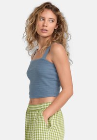 Modrý crop top s hrubými ramienkami a textúrovaným pleteným vzorom. Skombinovaný so zeleno-bielymi kockovanými nohavicami, ruky v vreckách.