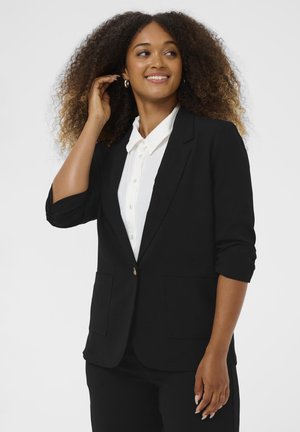 Donna con capelli ricci sorridente, che indossa un blazer nero sopra una camicia bianca con bottoni, con un braccio sollevato vicino alla testa.