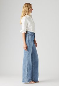 Jeans en denim bleu clair taille haute à jambe large, avec une finition nette et une texture légèrement délavée, associés à une chemise blanche à boutons.