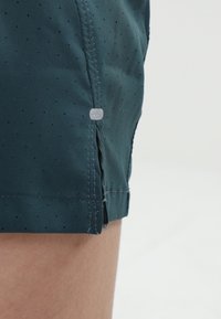 Tyrkysové šortky vyrobené z lehkého materiálu s perforovanou texturou, s bočními odvětrávacími otvory a malou průhlednou cedulkou poblíž lemování.