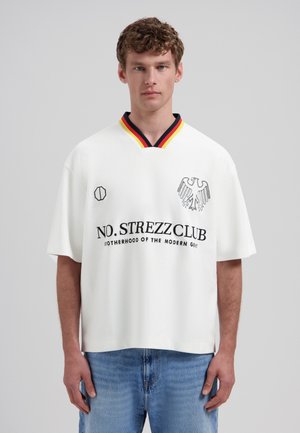 Ung mand med krøllet hår iført hvid oversized skjorte med sort ørneemblem, flerfarvet krave og sort tekst "NO.STREZZCLUB" kombineret med blå jeans.