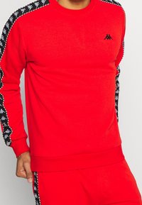 Sweat-shirt rouge avec un col rond, des poignets côtelés et des accents latéraux présentant un motif de logo répété en noir et blanc.