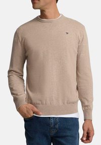Suéter de punto beige con mangas largas, escote redondo y puños acanalados. Presenta un pequeño logo en el lado izquierdo del pecho y un diseño ajustado.