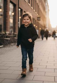 Dziecko w czarnej bluzie z zamkiem i niebieskich dżinsach idzie po brukowanej ulicy, ma na sobie beżowe buty, a w tle prześwieca światło słoneczne.