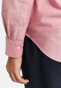Helles rosa Hemd mit langen Ärmeln, strukturiertem Stoff und weißen Knöpfen an den Manschetten. Getragen mit dunkelblauen Hosen, die die Länge des Hemdes betonen.