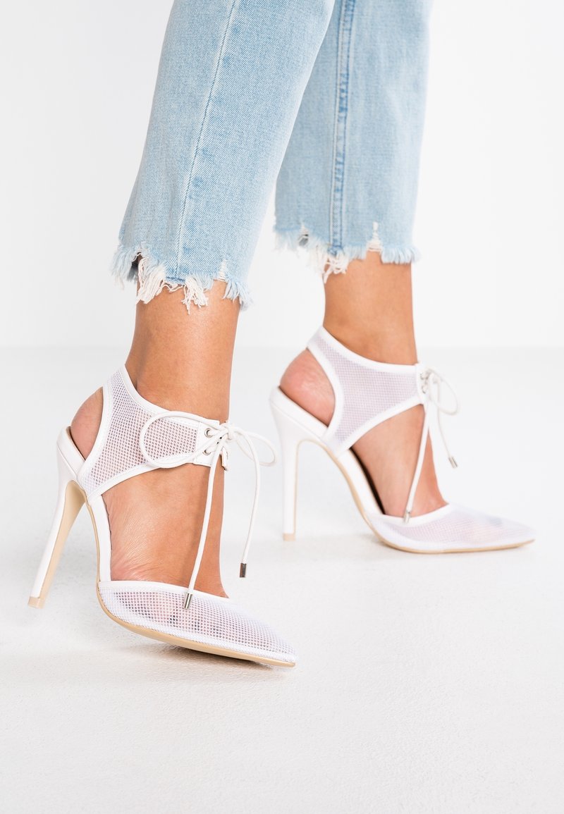 Chaussures à talons hauts en mesh blanc avec brides de cheville et lacets portées avec un jean bleu clair à ourlet effiloché sur une surface blanche.