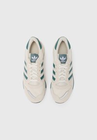 Beige sneakers met groene strepen, gemaakt van mesh en suede, met een ronde neus en witte accenten op de zool en de tong.