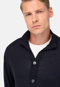 Marinblå cardigan med v-ringning och ribbad krage, med tre knappar, slät textur och en figurnära design. Bärs över en vit t-shirt.