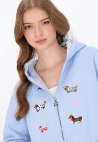 Hellblauer Kapuzenpullover mit Reißverschluss, bestickt mit Hundemotiven, Spitzenbesatz an der Kapuze und bunten Sternakzenten. Verfügt über einen dekorativen Reißverschlussanhänger.