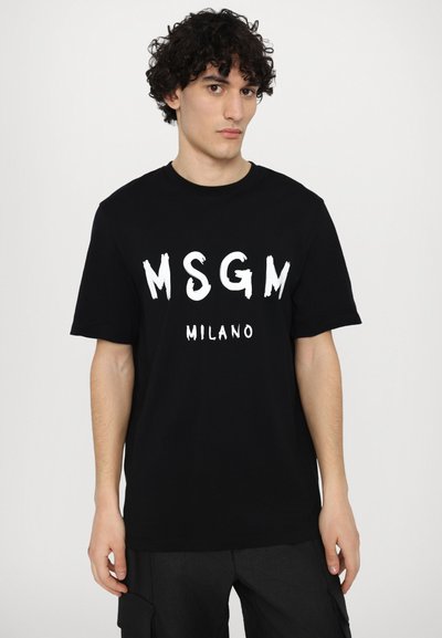 Musta puuvillatee, jonka edessä on valkoisella painatuksella teksti "MSGM MILANO" rohkealla, siveltimellä kirjoitetulla tyylillä. Lyhyet hihat, pyöreä pääntie.