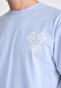 Helles blaues Baumwoll-T-Shirt mit weicher Textur, das ein weißes grafisches Design eines Gesichts, Flaschen und Blumen auf der Brust zeigt.